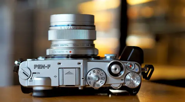 Silver Olympus PEN-F camera with a silver Olympus Digital 12mm 1:2.0 lens attached. The camera body features various dials and buttons, ready for capturing stunning images with its Olympus PEN F lenses.