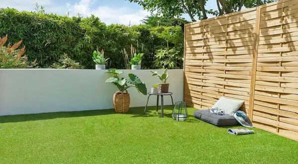 Un jardín moderno con césped artificial verde vibrante, rodeado de plantas y una valla de madera. Un rincón acogedor con cojines invita al descanso.
