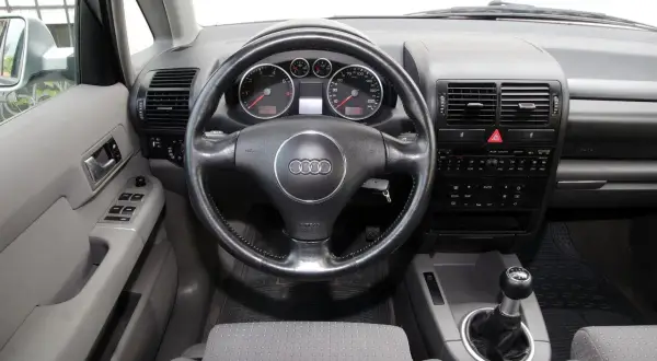 Wnętrze Audi A2 z kierownicą, deską rozdzielczą i dźwignią zmiany biegów. Widoczne wskaźniki, radio i przyciski.