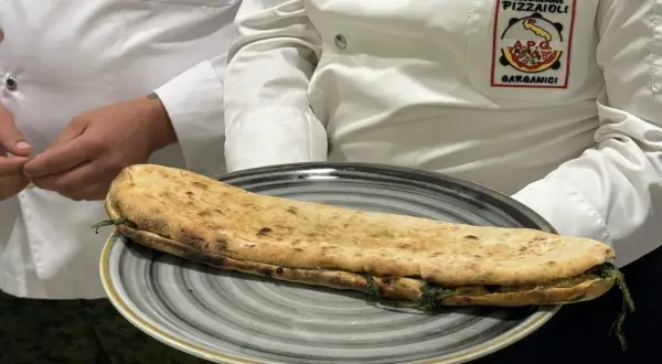 Una paposcia pugliese, ripiena e dorata, servita su un piatto grigio. Indossano giacche con stemmi dell'Associazione Pizzaioli Garganici.