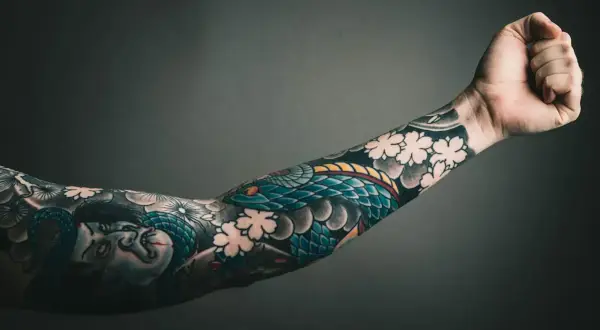 Ein Arm mit einem detailreichen, **heavy blackwork tattoo** von einer Schlange und einer Figur, umgeben von Kirschblüten.