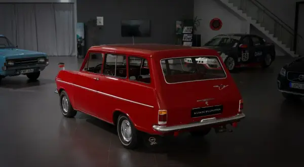 Röd veteranbil med ålder, en Opel Caravan, står i en bilhall bredvid en blå bil och en tävlingsbil.