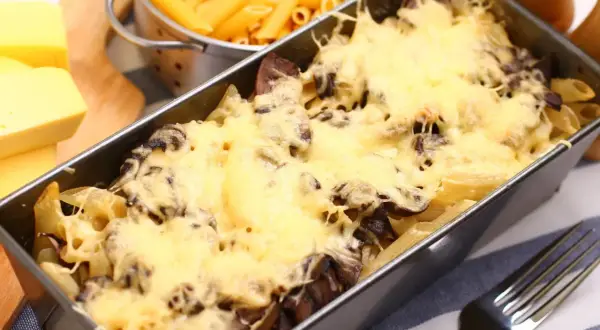 Pyszna zapiekanka z pieczarkami i makaronem penne, zapieczona pod złotym serem. Obok surowy makaron i kawałki sera.