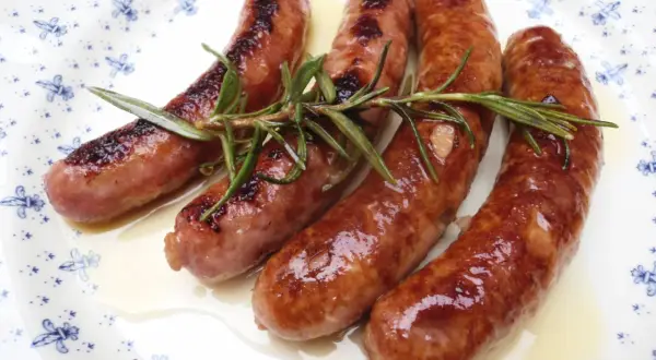 Salchichas doradas en salsa, con romero fresco, servidas en un plato blanco con diseño azul.