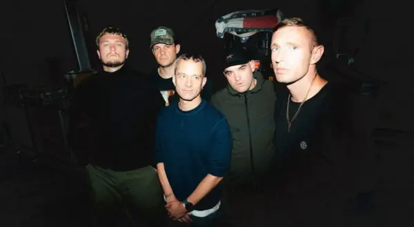 Five men stand in a dimly lit room, ready to perform. One holds a high vis setlist, hinting at the night's energetic show.