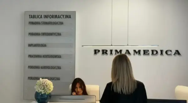 Recepcja kliniki PRIMAMEDICA z tablicą informacyjną i pracownicą.