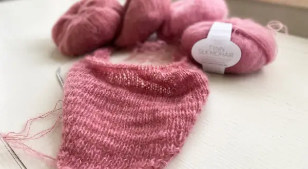 Zartrosa Mohair-Garn liegt neben einem Strickprojekt. Die feinen Fasern des Mohairs fusseln leicht und versprechen ein luftiges, weiches Ergebnis.
