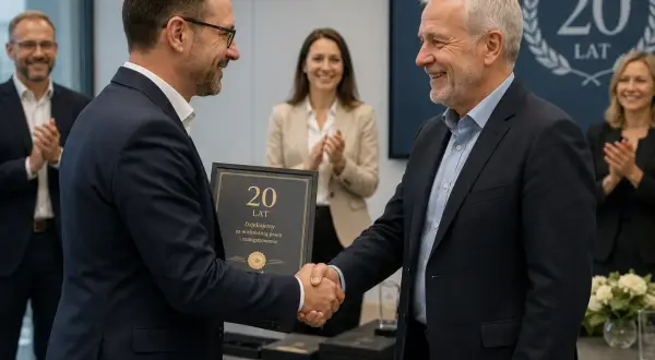 Dwóch mężczyzn w garniturach ściska sobie dłonie. Jeden z nich wręcza drugiemu ramkę z napisem "20 lat". W tle widać innych pracowników gratulujących jubilatom.