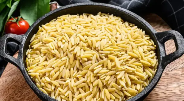 Makaron orzo w czarnej patelni, obok pomidorów i ziół.