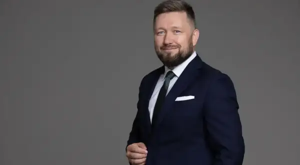 Mężczyzna w granatowym garniturze i zielonym krawacie, uśmiecha się na szarym tle.