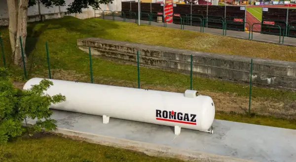 Biały zbiornik gazu ROMGAZ stoi na betonowej podstawie. Widać ogrodzenie i trawę, a w tle trybuny stadionu. Odległość gazociągu od ogrodzenia jest widoczna.