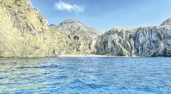 Aguas azules bañan la costa rocosa de una cala en Almería. Montañas escarpadas y una pequeña playa de arena componen el paisaje.