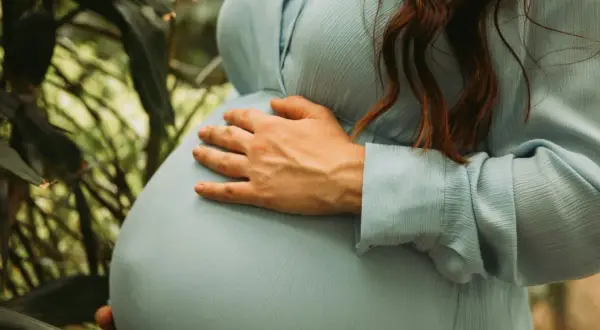 Gravid i vecka 5, en kvinna med ljusblå klänning håller om sin mage bland gröna blad.