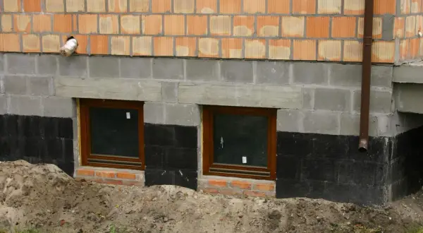 Prace przy izolacji fundamentów w starym domu. Dwa okna piwniczne, czarna izolacja przeciwwilgociowa i nowe mury.