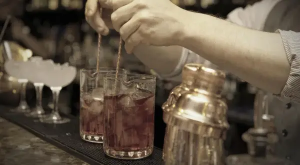 Barkeeper mixt einen roten Bier Cocktail mit Eis in einem Glas. Daneben steht ein goldener Shaker.