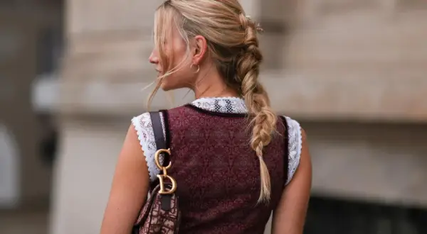 Blonde Zopf-Frisur mit Blumenakzenten, perfekt für Oktoberfest Frisuren. Trägt Dirndl und eine Designer-Tasche.
