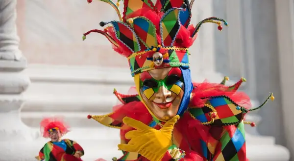 Un personaggio con un elaborato tipo di maschera di carnevale, colorato e festoso, con un cappello a punta e guanti gialli.