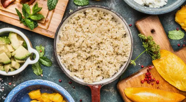 Perfekt gekochtes Quinoa in einer Pfanne, umgeben von frischen Zutaten wie Avocado, Mango und Tomaten.