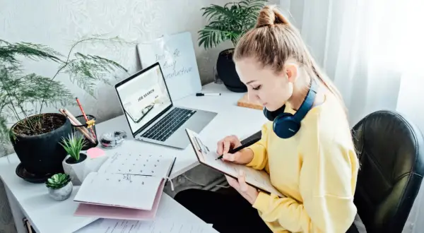 Kobieta pracuje nad CV, szukając pracy. Na biurku laptop z napisem "Job Search", kalendarz i notatnik.