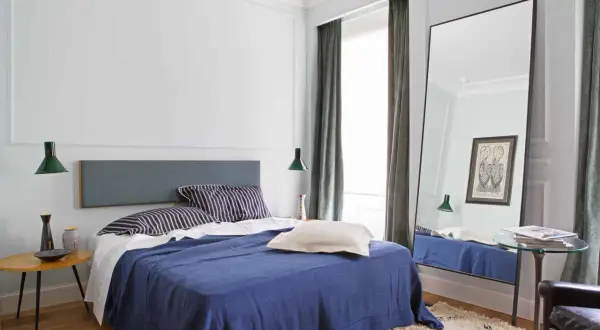 Modern mens bedroom ideas: a navy blue bed with striped pillows, a grey headboard, and a large floor mirror reflecting the room.