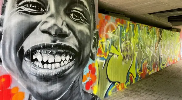 Graffiti-Porträt eines lachenden Kindes neben einer bunten Wand mit Schriftzügen.