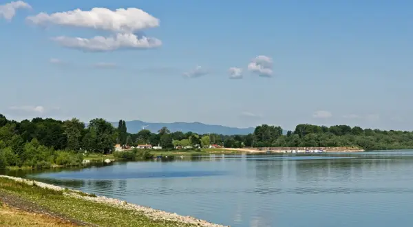 Spokojne jezioro otmuchowskie z piaszczystą plażą, otoczone zielenią. W oddali widać góry i wiatraki.