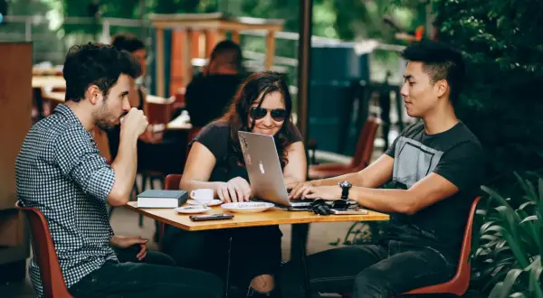 Tres jóvenes colaboran en un portátil en una cafetería al aire libre, un ejemplo de cómo ser nómada digital.