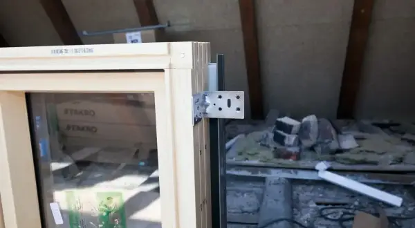 Drewniana rama okna z metalowym wspornikiem, gotowa do montażu. W tle widać materiały budowlane i belki dachu. Okno jest wykonane z materiału podobnego do **pvc**.