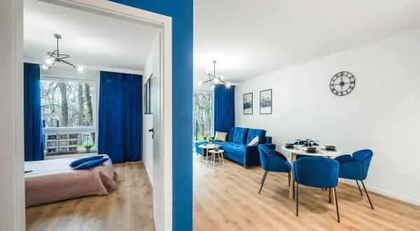 Nowoczesny pokój nad morzem z niebieskimi akcentami. Widok na salon z sofą i jadalnią oraz sypialnię z łóżkiem.