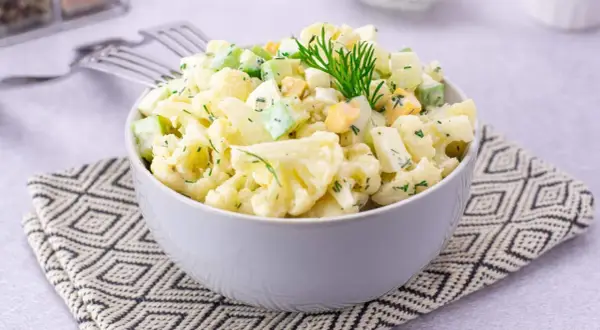 Sałatka z kalafiorem, jajkiem i ogórkiem, udekorowana koperkiem, podana w misce na wzorzystej serwetce.