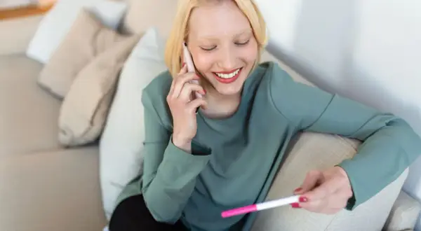 Uśmiechnięta kobieta z testem ciążowym w dłoni rozmawia przez telefon, zastanawiając się, kiedy powiedzieć pracodawcy o ciąży.