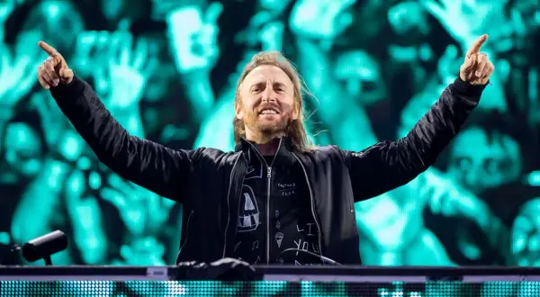 David Guetta na jednym ze swoich koncertów, z rękami uniesionymi w geście radości. Tłum na tle ekranów LED.