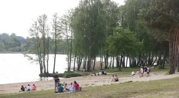 Ludzie odpoczywają na piaszczystej plaży nad jeziorem Pławniowice, ciesząc się wiosennym dniem wśród drzew.