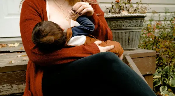 En mamma ammar sitt barn utomhus. Hon bär en röd kofta och mörka byxor. Bilden ger en känsla av lugn och närhet, och kan inspirera till att gå ner i vikt amning på ett hälsosamt sätt.