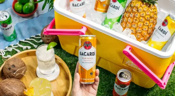 Letnia piknikowa atmosfera z drinkami Bacardi. W żółtej lodówce schłodzone puszki rumowych koktajli i ananas. Na trawie dwa drinki, kokosy i limonki.