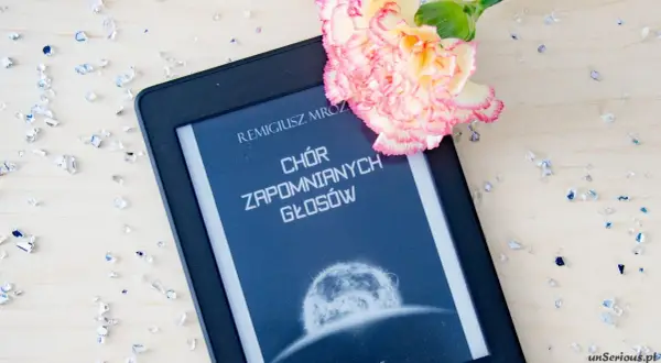 Elektroniczna książka z tytułem "Chór zapomnianych głosów" Remigiusza Mroza, ozdobiona kwiatem i rozsypanymi kryształkami.