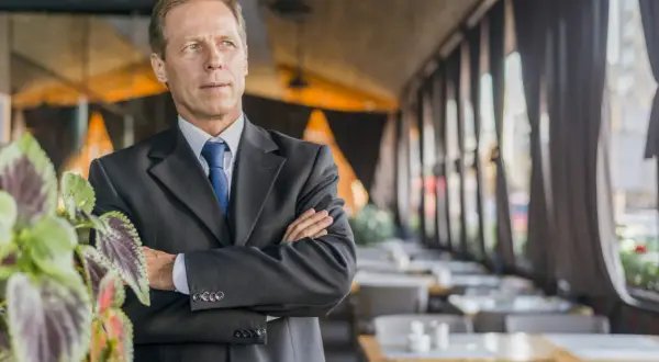 Uomo d'affari con braccia conserte, un chief people officer che guarda lontano, con piante verdi in primo piano e un ristorante sullo sfondo.