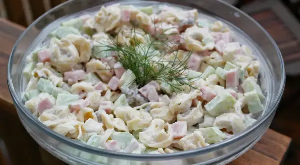 Pyszna sałatka z tortellini, szynką i ogórkiem, udekorowana gałązką koperku. Idealna na letnie przyjęcie.