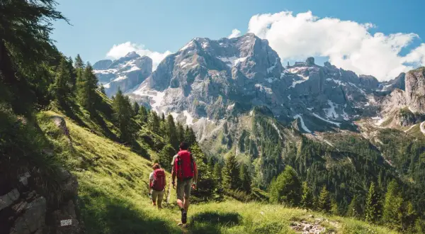 Dos personas disfrutan del senderismo y trekking por un sendero verde con imponentes montañas nevadas de fondo.