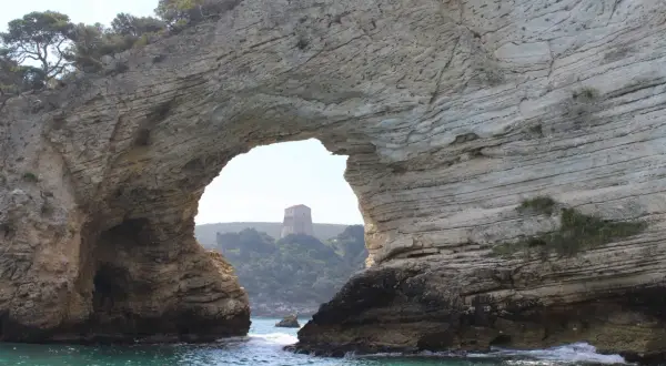 Arco naturale di roccia che incornicia una torre sulla collina, vista dalla baia San Felice Vieste. Acque turchesi e vegetazione mediterranea.