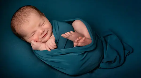 Un bébé endormi, emmailloté dans un tissu bleu canard, sourit paisiblement. Une belle image pour célébrer une naissance, un moment de joie et de gratitude, que dire pour une naissance islam.