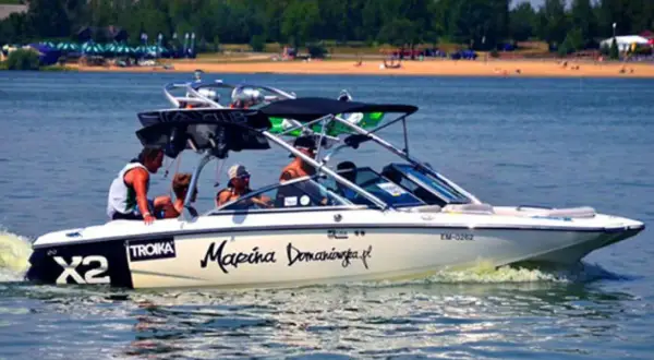 Łódź motorowa z pasażerami płynie po wodzie, tworząc pianę. Na burcie widnieje napis "Marina Domaniewska".
