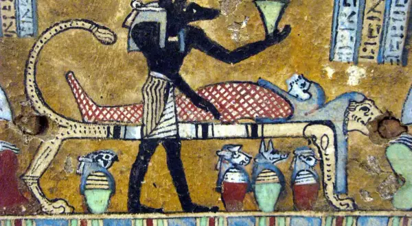 Anubis, bóg z głową szakala, stoi przy katafalku z mumią. W ręku trzyma naczynie, a obok stoją urny z głowami zwierząt.