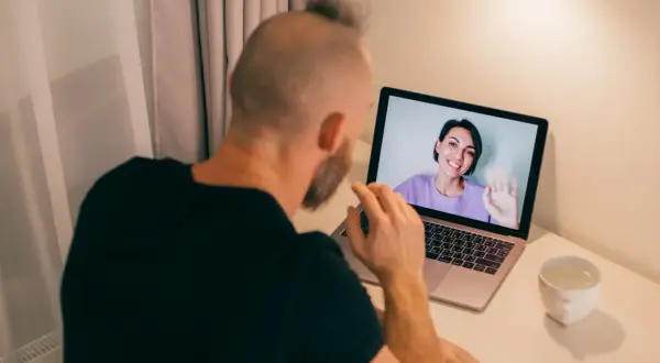 Mężczyzna odbywa konsultację psychologiczną online. Na ekranie laptopa widoczna uśmiechnięta kobieta machająca ręką.