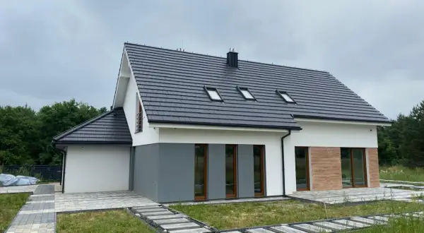 Nowy dom z szarym dachem i białymi ścianami, gotowy do odbioru. Wokół ścieżki z kostki brukowej i trawa.