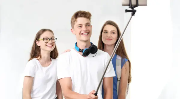 Trzech przyjaciół robi sobie selfie z kijkiem. Chłopak z przodu uśmiecha się szeroko, dziewczyna w okularach po lewej też, a ta z tyłu patrzy w obiektyw. To prosty sposób, jak zrobić ładne zdjęcie grupowe.