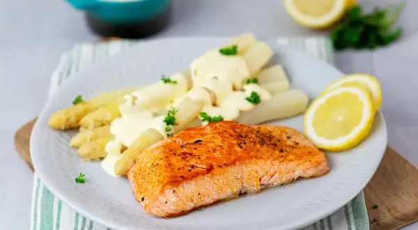 Ein saftiges Stück Lachs liegt neben weißem Spargel mit Sauce Hollandaise auf einem Teller. Garniert mit Petersilie und Zitronenscheiben.