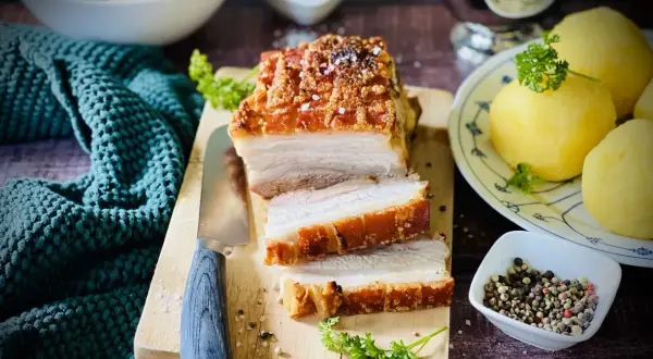 Ein saftiger Schweinebraten mit knuspriger Kruste, auf einem Holzbrett angeschnitten, daneben Klöße und Soße.