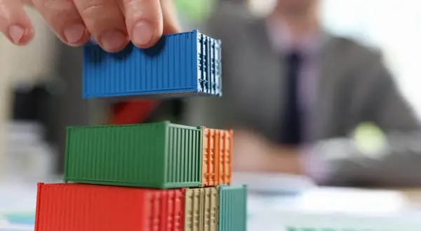 Dłoń układa niebieski kontener na stosie kolorowych modeli. Analiza kosztów transportu i potencjalnego cła.