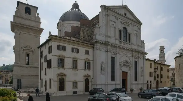 La piazza di San Giorgio a Verona, con la chiesa imponente, la torre campanaria e alcune persone che passeggiano tra le auto parcheggiate.
